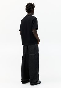 Polo shirt nera a maniche corte abbinata a pantaloni in denim scuri a gamba larga; cuciture visibili e grandi tasche posteriori. Il modello è di spalle.