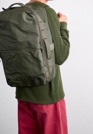 Personne portant une chemise verte à manches longues et un pantalon rouge, portant un grand sac à dos vert olive sur le dos, sur un fond blanc uni.