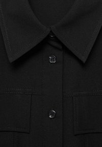 Chemise noire à col avec un devant à boutons, en tissu texturé et dotée de deux poches poitrine. Détails en boutons noirs et coutures contrastées.