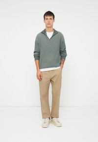 Pulover verde cu model canelat, cu guler, tricou alb dedesubt, pantaloni bej cu croială dreaptă și adidași crem. Design simplu, cu o croială lejeră.