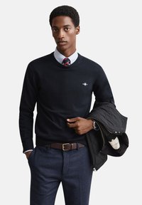 Tamsiai mėlynas megztinis su apvalia kaklo iškirpte, ažūrinėmis rankogaliais ir mažu logotipu. Derinamas su šviesiai mėlyna marškinių apykakle ir tamsiai mėlynomis tekstūruotomis kelnėmis.