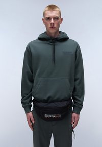 Sudadera verde con bolsillo frontal y cordón. Riñonera negra con logo, llevada en la cintura. El modelo está de pie frente a un fondo neutro.