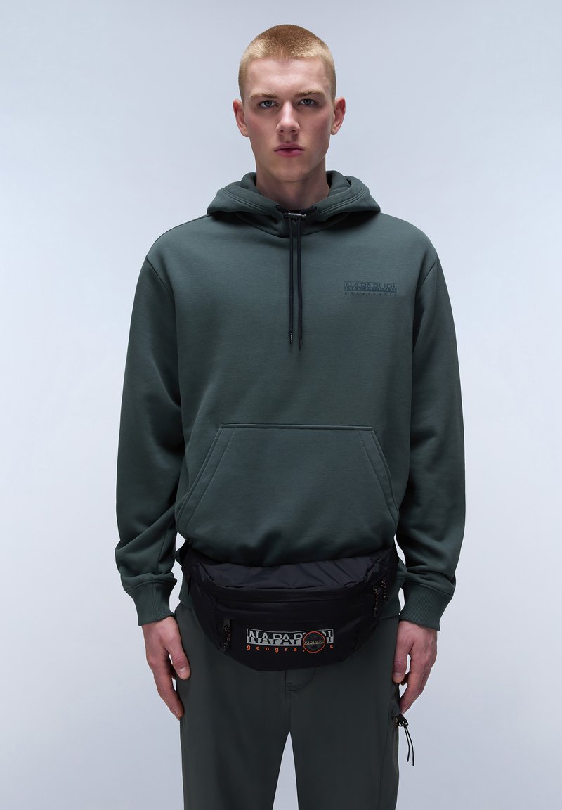 Sudadera verde con bolsillo frontal y cordón. Riñonera negra con logo, llevada en la cintura. El modelo está de pie frente a un fondo neutro.