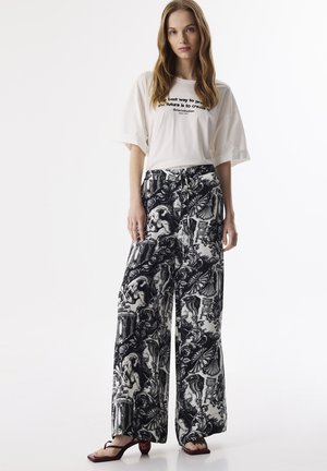 Witte T-shirt met zwarte tekst, gecombineerd met wijde broek met een zwart-wit ornamenteel patroon en platte zwarte sandalen.