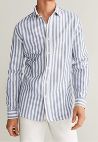 Muž ve modro-bílém pruhovaném dlouhém tričku s knoflíky a bílých kalhotách, stojící na neutrálním pozadí.