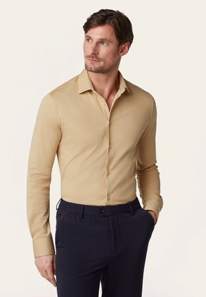 Man draagt een beige overhemd met lange mouwen en knopen en donkerblauwe broek, kijkt iets naar links, hand in zijn zak.