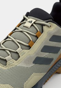 Chaussure de sport gris clair avec tige en maille, accents texturés, rayures gris foncé et détails de laçage orange. Semelle en caoutchouc visible en bas.