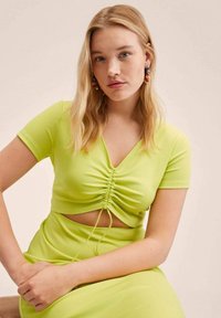 Jeune femme aux cheveux blonds portant un crop top froncé vert citron et une jupe, boucles d'oreilles colorées en forme d'anneau, assise les mains jointes.