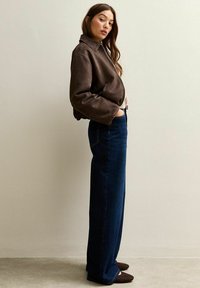 Chaqueta bomber de cuero marrón con cierre de cremallera, combinada con unos pantalones vaqueros de talle alto de pierna ancha azul oscuro y zapatos de gamuza marrón.