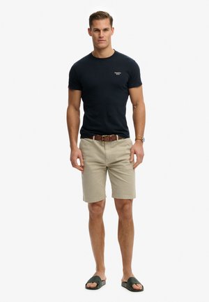 Homme debout, portant un t-shirt noir à manches courtes, un short beige au genou avec une ceinture marron, des sandales noires à enfiler et une montre-bracelet.