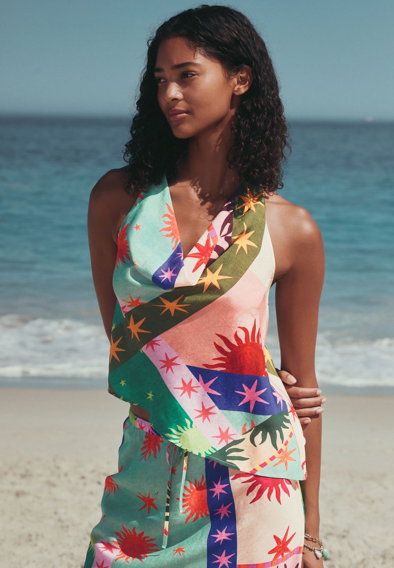 Femme aux cheveux bouclés debout sur une plage, portant un haut sans manches coloré à motifs de soleil et d’étoiles et un short assorti.