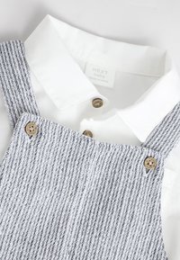 Chemise bébé blanche à col sous une salopette tricotée grise et blanche à rayures avec des boutons en bois portant la marque "next".