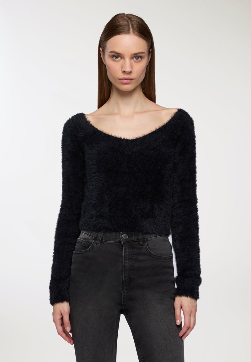 Donna con capelli castani lisci, che indossa un maglione nero morbido a spalle scoperte e jeans scuri a vita alta, in piedi contro uno sfondo bianco.
