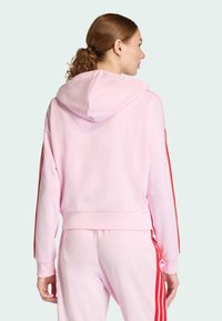 Ružová mikina s kapucňou a červenými bočnými pruhmi, voľný strih a rebrované manžety. Mäkký materiál a skrátený dizajn. Viditeľné detaily šitia.