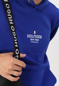 Sudadera con capucha de color azul royal con un bolsillo frontal, que presenta un logo blanco "HILFIGER NEW YORK" y un pequeño acento tricolor. Correa negra visible.
