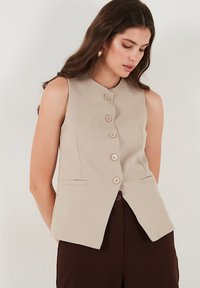 Beige ujjatlan mellény, kerek nyakkivágással, hat gombbal és két elülső zsebbel, sima anyagból készült.