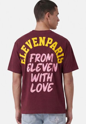 Hombre de espaldas que lleva una camiseta burdeos con texto amarillo "ELEVENPARIS" y texto rosa "FROM ELEVEN WITH LOVE" en la espalda.