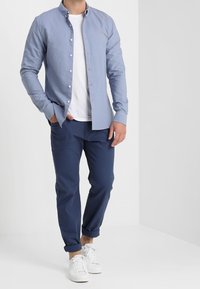 Chemise à manches longues bleu clair avec boutons, portée sur un t-shirt blanc, associée à un pantalon bleu marine et des baskets blanches. Design simple, coupe décontractée.