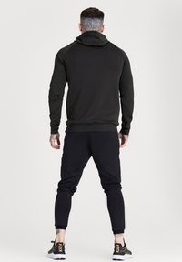 Felpa sportiva nera con un design aderente, dotata di cappuccio e maniche lunghe. Indossata con pantaloni jogger aderenti abbinati e sneaker nere.