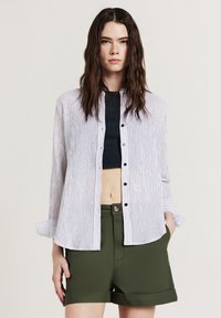 Gestreiftes, weißes und marineblaues Hemd mit Knopfleiste, Kragen und langen Ärmeln, kombiniert mit olivgrünen Shorts, die eine lässige Passform und ein leichtes Material zeigen.