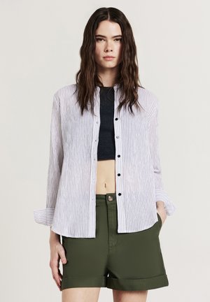 Camicia a righe bianca e blu navy con colletto e maniche lunghe, abbinata a pantaloni corti verde oliva, che mostrano una vestibilità rilassata e un tessuto leggero.