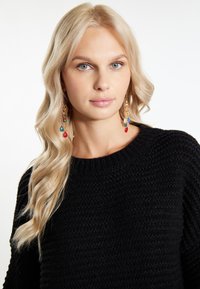 Maglione nero lavorato a maglia con una finitura testurizzata, caratterizzato da un scollo ampio. Il modello indossa orecchini lunghi e colorati con perline e pendenti.