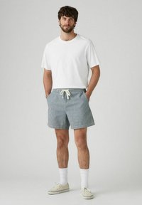 T-shirt bianca a maniche corte abbinata a pantaloni da bagno blu a righe con vita regolabile, calzini bianchi e scarpe da ginnastica chiare.