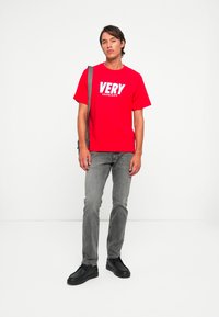 Vyras, apsivilkęs raudoną marškinėlius "VERY Calvin Klein", pilkus džinsus, juodus batus ir nešantis pilką petinę kuprinę, stovi prieš baltą foną.