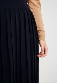 Gonna plissettata navy realizzata in tessuto testurizzato, dotata di un girovita aderente e morbide pieghe cadenti, abbinata a un maglione beige a maniche lunghe.