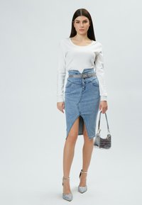 Jupe crayon en denim taille haute, avec fente à l'avant et fermeture à bouton. Associée à un haut blanc à manches longues et des chaussures à talons argentées.