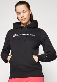 Černá mikina s kapucí vyrobená z bavlněné směsi, která má velké vyšívané logo "Champion" v bílé barvě s červeným akcentem, přední klokankovou kapsu a ribbované manžety.