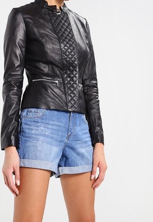 Vrouw draagt een zwart gewatteerd leren jack met ritssluitingen en blauwe opgerolde spijkershorts, staand tegen een effen achtergrond.