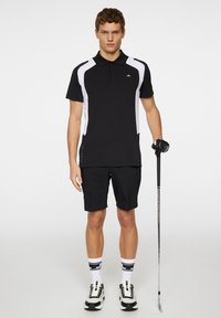 Svart pikétröja med vita accenter, tillsammans med svarta shorts. Tillbehör inkluderar en golfhandske, klubba och mönstrade strumpor med sneakers.