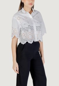 Camicetta bianca cropped con intagli floreali a occhio e orlo scallop. Presenta un colletto e maniche corte. Abbinata a pantaloni neri.