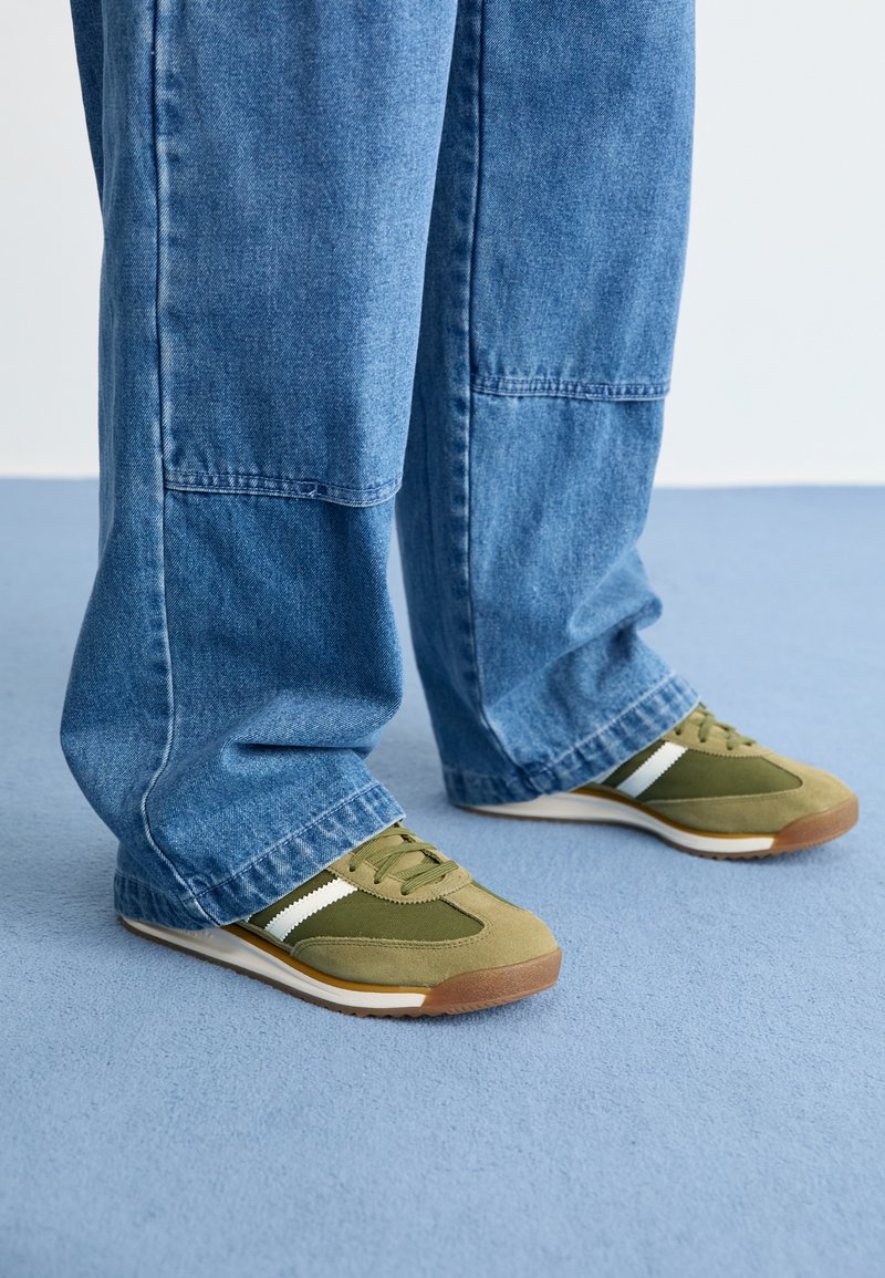 Sapatilhas verdes e bege com detalhes brancos numa sola off-white, combinadas com jeans de ganga azul soltos. A superfície é texturada e o corte é descontraído.
