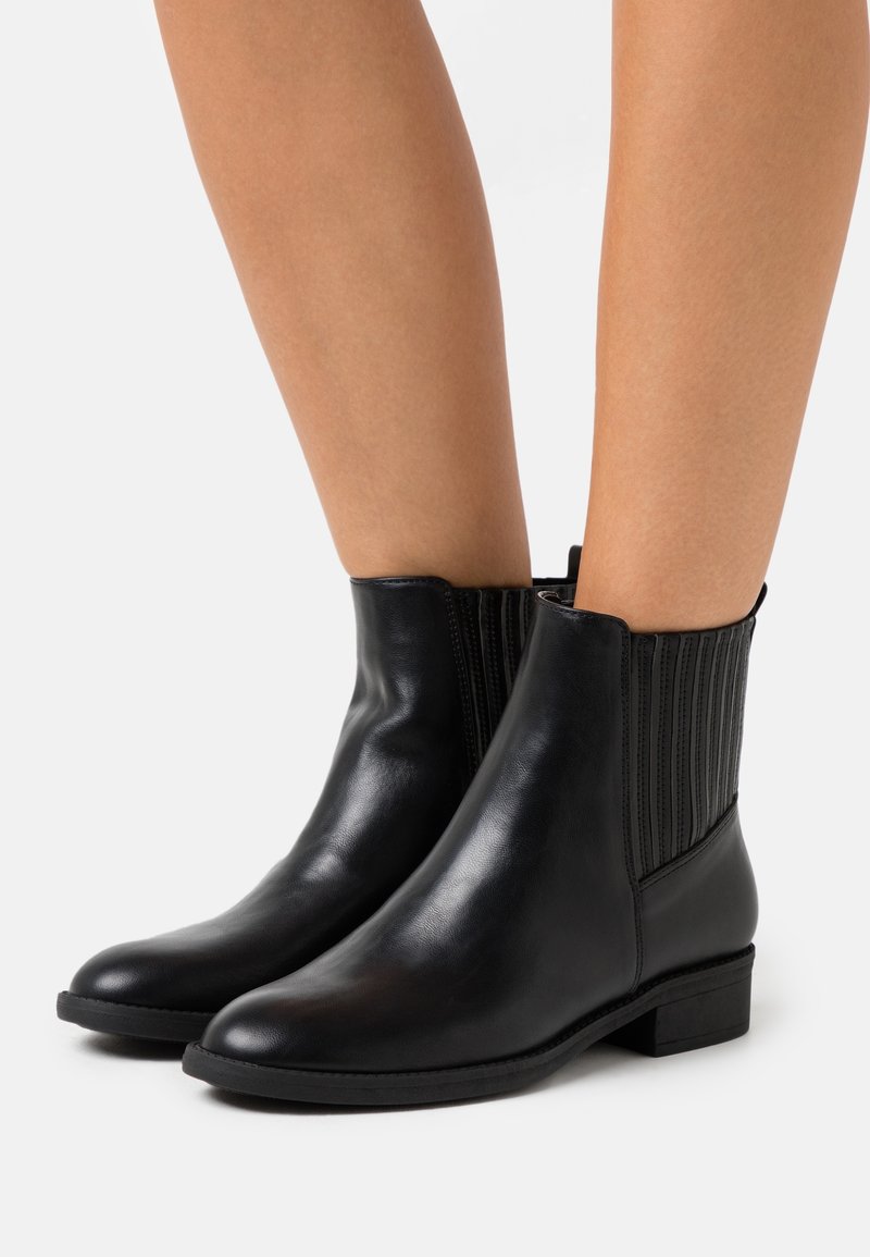 Bottines en cuir noir avec panneaux latéraux côtelés élastiques portées sur jambes nues sur un fond clair uni.