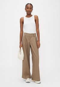 Sieviete stāv, tērpusies baltā bezrokasnītei, brūnās augsta vidukļa platajās biksēs, baltos sporta apavos un rokās tur baltu rokassomu.