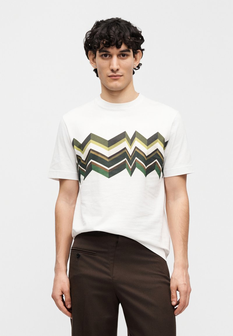 White cotton t-shirt featuring a zigzag pattern in green and olive hues across the chest, paired with brown trousers.
