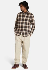 Camisa de cuadros marrón y beige, diseño de botones, usada por fuera sobre pantalones beige claros, combinada con zapatos marrones de cordones.