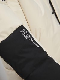Puffjack met een crèmekleurig lichaam en zwarte mouwen. Glad oppervlak met logo-details in het wit. Zichtbare stiksels en knoopaccenten.