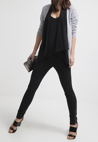 Schwarzes ärmelloses Oberteil mit grauem offenen Cardigan, schmale schwarze Hose mit Reißverschlüssen an den Knöcheln und schwarze Sandalen, die eine strukturierte Clutch halten.