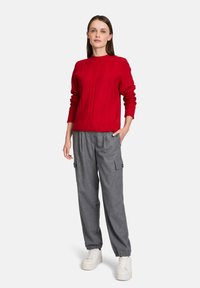 Maglione rosso a maglia grossa con maniche lunghe, abbinato a pantaloni cargo grigi con tasche. Il modello indossa sneaker bianche. Outfit semplice e casual.