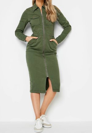 Vrouw draagt een olijfgroene jurk met lange mouwen en een ritssluiting, met voorzakken en witte sneakers, staand met handen in de zakken.