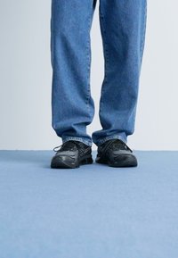 Svarta sportskor med texturerade ovandelar och snörning, bärs med löst sittande blå denimjeans, stående på en ljusblå yta.