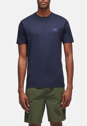 Uomo che indossa una t-shirt blu navy a maniche corte con piccolo logo e pantaloncini verde oliva fino al ginocchio, in piedi su sfondo neutro.