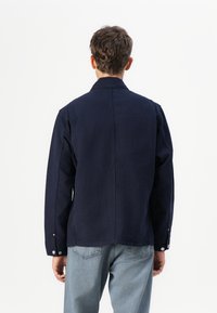 Chaqueta de color azul marino con cuello puntiagudo, mangas largas, costuras laterales y botones a presión en los puños. Tejido de algodón, diseño estructurado con corte recto.