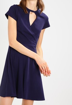 Kobieta ubrana w granatową sukienkę z krótkim rękawem, z dekoltem w kształcie łezki i rozkloszowanym dołem, stojąca z rękami złączonymi przed sobą.