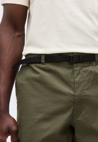 Άτομο με σκουρόχρωμο δέρμα που φοράει χακί παντελόνι με μαύρη ζώνη και ένα ανοιχτό μπεζ t-shirt σκουπισμένο μέσα.