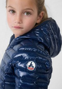 Marineblauwe gewatteerde jas met een glanzende afwerking, voorzien van horizontale doorgestikte details en een ronde JOTT-logo op de linkermouw.