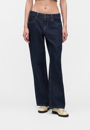 Femme portant un jean bleu foncé à jambes larges et des chaussures à lacets couleur crème, debout devant un fond uni.